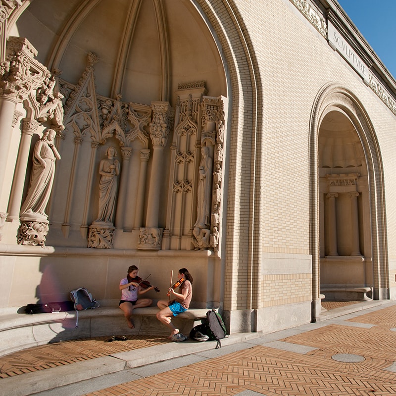 Photo of a campus building with two students playing violin